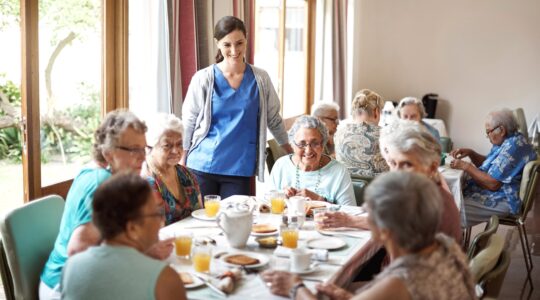 Carer with elderly in aged care home dining room. | Newsreel