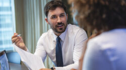 Business man talking to a business woman while holding a document. He is happy and smiling. He is wearing a tie.