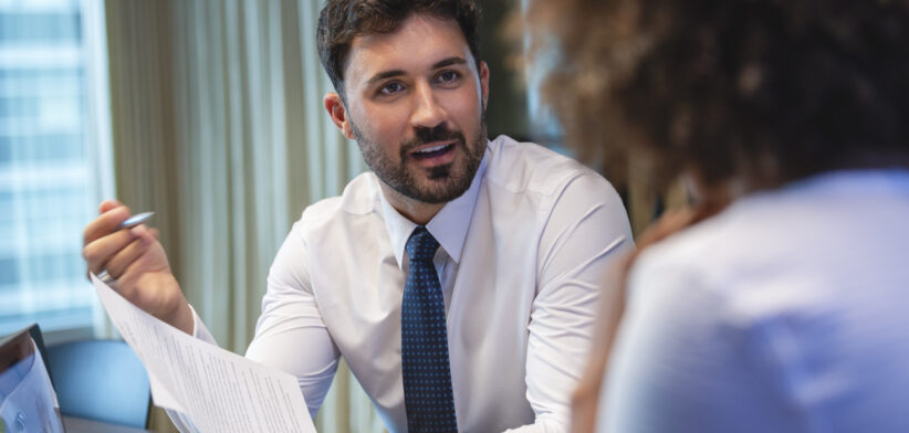 Business man talking to a business woman while holding a document. He is happy and smiling. He is wearing a tie.