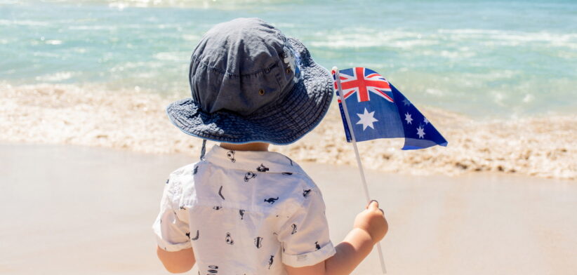 Little boy wearing hat holding Australian flag on a sandy ocean beach. Australia Day concept