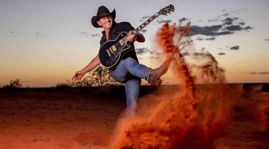 Man with guitar kicking up red dust in bush - Newsreel