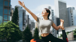 Athletic young Asian woman practicing yoga outdoors in city park against urban cityscape in the morning