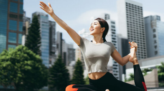 Athletic young Asian woman practicing yoga outdoors in city park against urban cityscape in the morning