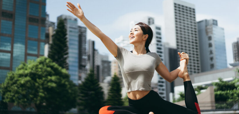 Athletic young Asian woman practicing yoga outdoors in city park against urban cityscape in the morning