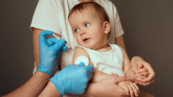 A caregiver holds a toddler in preparation for a vaccination. The medical professional, wearing gloves, is ready to administer a shot while the child curiously observes