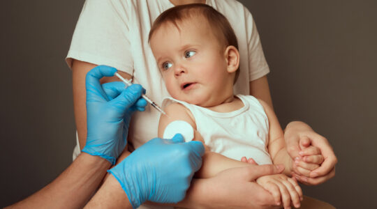 A caregiver holds a toddler in preparation for a vaccination. The medical professional, wearing gloves, is ready to administer a shot while the child curiously observes