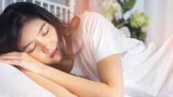 Woman stretching in bed after waking up, back view. Woman sitting near the big white window while stretching on bed after waking up with sunrise at morning, back view.