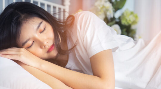 Woman stretching in bed after waking up, back view. Woman sitting near the big white window while stretching on bed after waking up with sunrise at morning, back view.