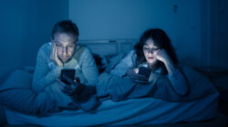 Life style portrait of young bored couple in bed at night on smart phones obsessed with games, social media, apps ignoring each other. Relationship communication problems and phone addiction concept.