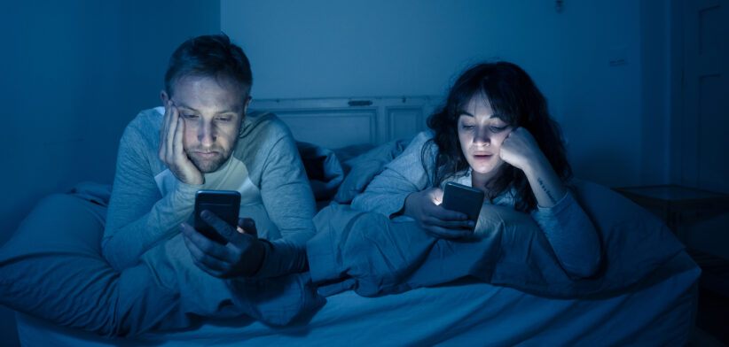 Life style portrait of young bored couple in bed at night on smart phones obsessed with games, social media, apps ignoring each other. Relationship communication problems and phone addiction concept.