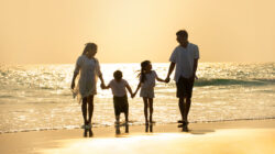 Happy Family parents with two child boy and girl holding hands and walking together on the beach at summer sunset. Father, mother and kids enjoy and having fun in outdoor holiday vacation.