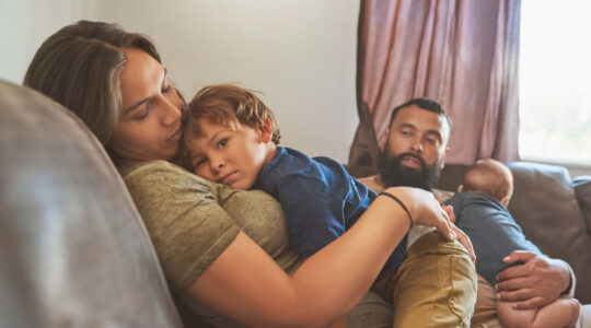 Shot of a little boy resting on his mother’s chest with his baby brother and father in the background