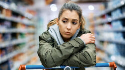Young sad woman leaning on shopping cart while standing among produce aisle at supermarket.