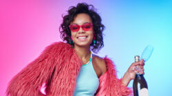 Beautiful African woman in trendy clothes holding bottle with sparkling wine against colorful background