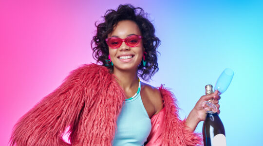 Beautiful African woman in trendy clothes holding bottle with sparkling wine against colorful background