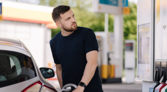 Bearded man refuelling car on gas station and looking into his smartphone. Man compares fuel prices