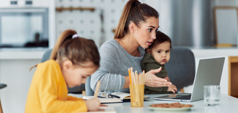 A busy working mother having a work-related video call at home on her laptop and looking after her children