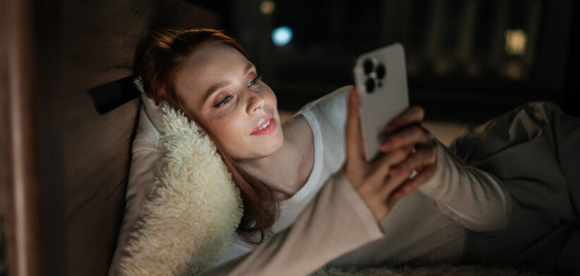 Cute redhead young woman lying in bed at night, face illuminated by soft glow of smartphone, smiling scrolling social media content and enjoying moment of connection