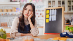 hardworking bakery owner focuses on her laptop inside the cake shop, her face showing stress and determination as she manages orders and business tasks. Nearby, a dedicated male baker and a thoughtful Korean barista work diligently reflecting challenge