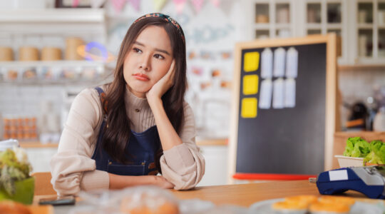 hardworking bakery owner focuses on her laptop inside the cake shop, her face showing stress and determination as she manages orders and business tasks. Nearby, a dedicated male baker and a thoughtful Korean barista work diligently reflecting challenge