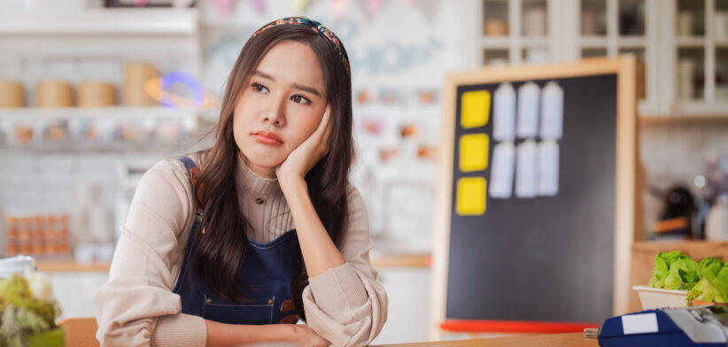 hardworking bakery owner focuses on her laptop inside the cake shop, her face showing stress and determination as she manages orders and business tasks. Nearby, a dedicated male baker and a thoughtful Korean barista work diligently reflecting challenge