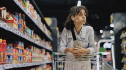 Young woman shopping in a supermarket, pushing a shopping cart through the aisles while browsing and selecting products. She is stocking up on weekly groceries, household cleaning supplies, and personal care items.