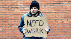 Homeless holds piece of cardboard with inscription Need Work. Refugee is at brick wall looking for job. Male tramp in dirty clothes. Below poverty line. Bearded fugitive beggar. Immigrant. Drunkenness