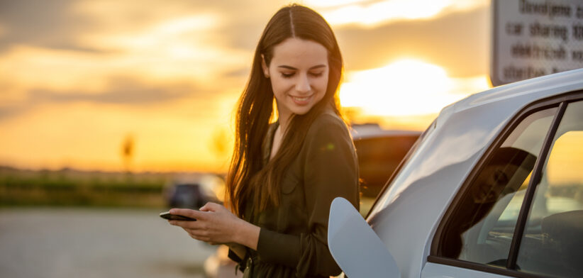 Woman waiting for electric car to charge in the parking lot at sunset