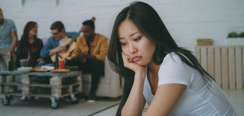 Young asian girl feels upset and isolated while her flatmates celebrating party at home indoors