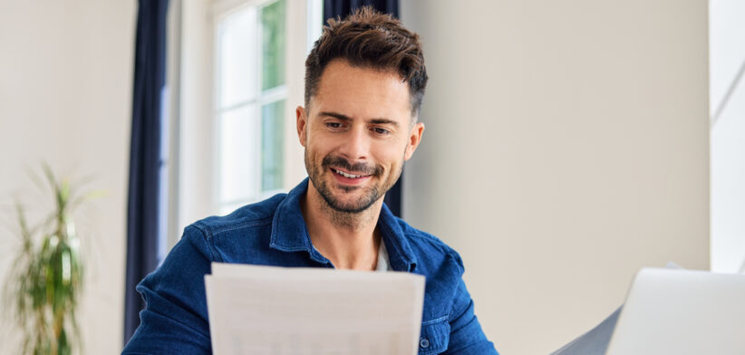 Happy man looking at document while working on laptop from home at living room