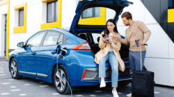Young friends charging automobile battery from public station and standing near parking for electric car. Man and woman talking and drinking coffee in city