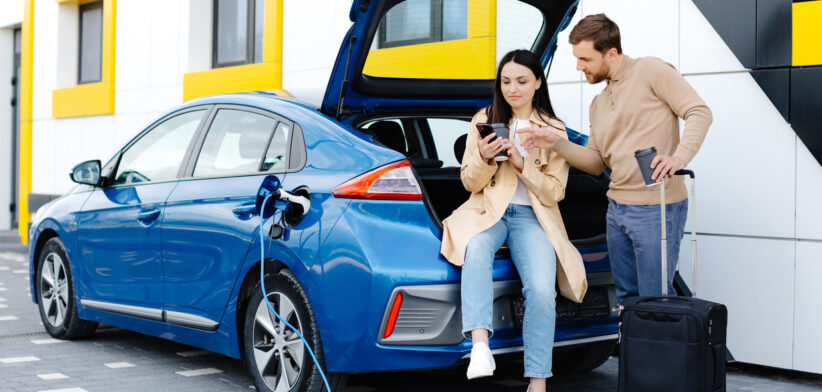 Young friends charging automobile battery from public station and standing near parking for electric car. Man and woman talking and drinking coffee in city