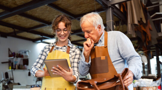 Senior artisan and apprentice crafting leather handbag in a workshop together