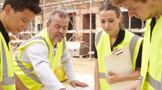 Builder On Building Site Discussing Work With Apprentice