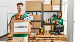 Young hispanic volunteer couple working at charity center. Man smiling happy holding box with toys to donate.