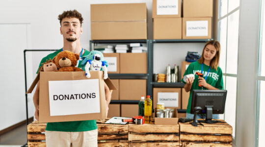 Young hispanic volunteer couple working at charity center. Man smiling happy holding box with toys to donate.