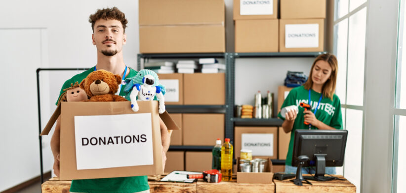 Young hispanic volunteer couple working at charity center. Man smiling happy holding box with toys to donate.