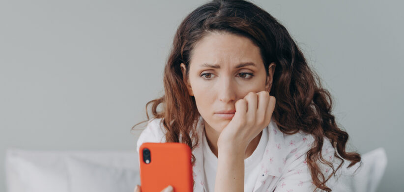 Stressed caucasian woman is doom scrolling. Girl reading morning news on smartphone in her bedroom.