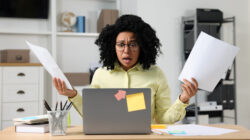 Deadline concept. Scared woman holding documents and looking at laptop in office