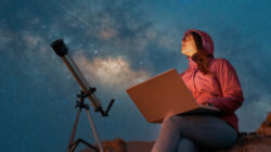 portrait woman sitting in the desert with a laptop next to a telescope at night watching the starry sky, astronomy and stargazing concept