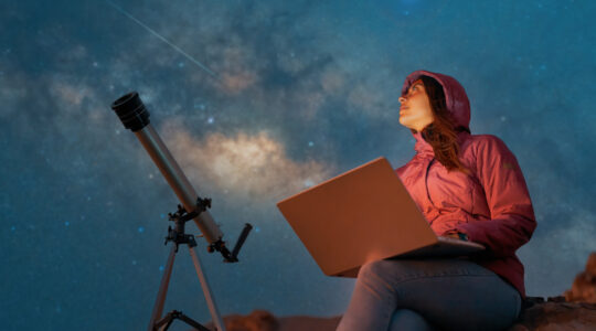 portrait woman sitting in the desert with a laptop next to a telescope at night watching the starry sky, astronomy and stargazing concept