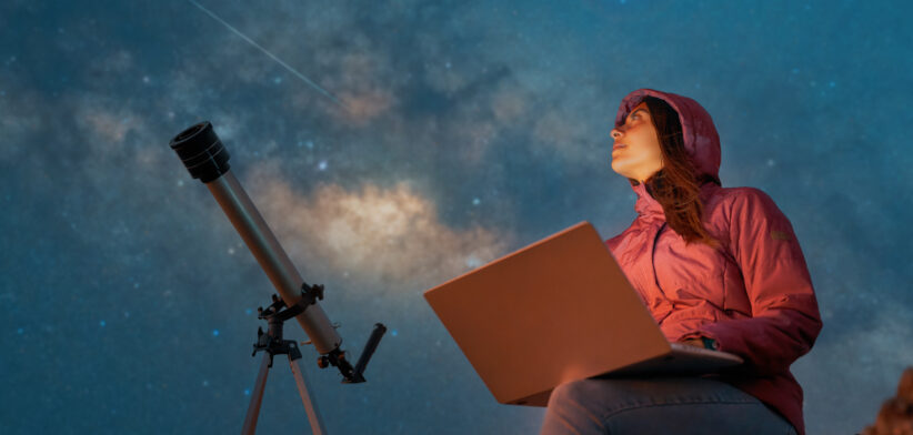 portrait woman sitting in the desert with a laptop next to a telescope at night watching the starry sky, astronomy and stargazing concept
