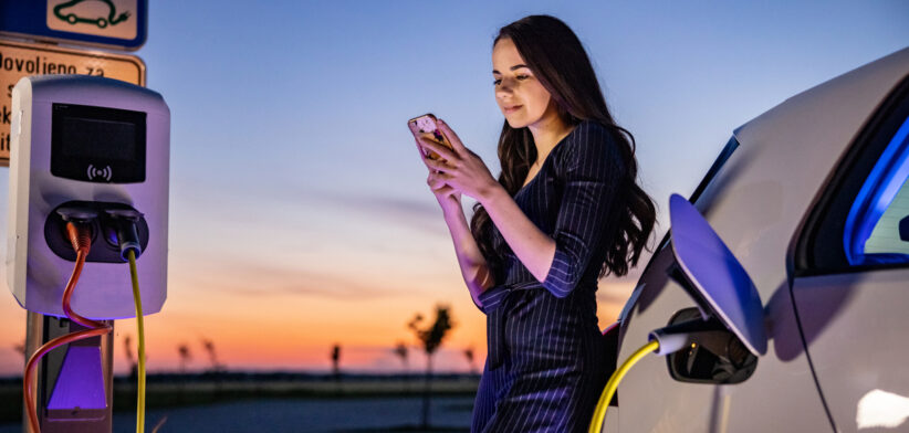 Young woman text messaging on mobile phone while waiting for electric car to charge