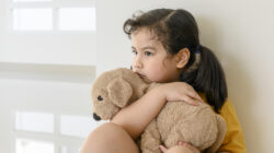 Little Asian girl stays home bored by school closings due to covid pandemic. Preschool girl looking outside her home window.
