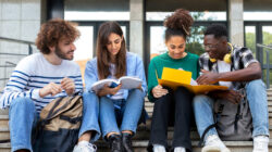 Happy diverse university student friends doing homework together sitting on campus steps.