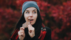 Young happy girl in a gray hat, a plaid scarf and a dark coat eats milk chocolate with whole nuts and smiles. Fall season. The concept of dealing with depression.
