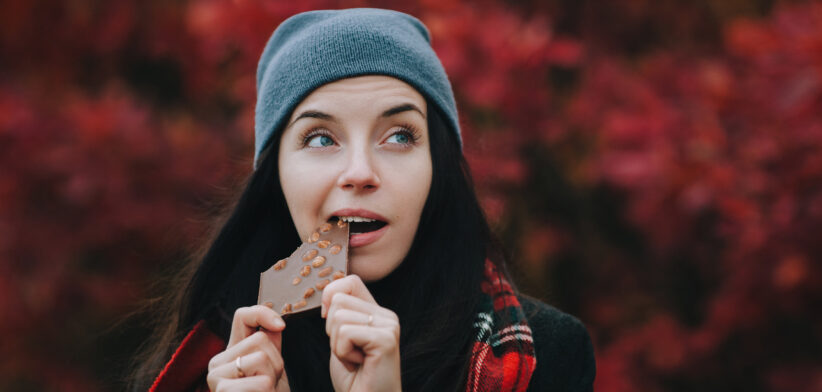 Young happy girl in a gray hat, a plaid scarf and a dark coat eats milk chocolate with whole nuts and smiles. Fall season. The concept of dealing with depression.