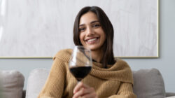Pretty, attractive, stylish young woman sitting on couch having raised glass with red wine, examine, taste beverage