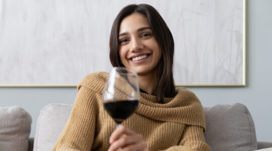 Pretty, attractive, stylish young woman sitting on couch having raised glass with red wine, examine, taste beverage