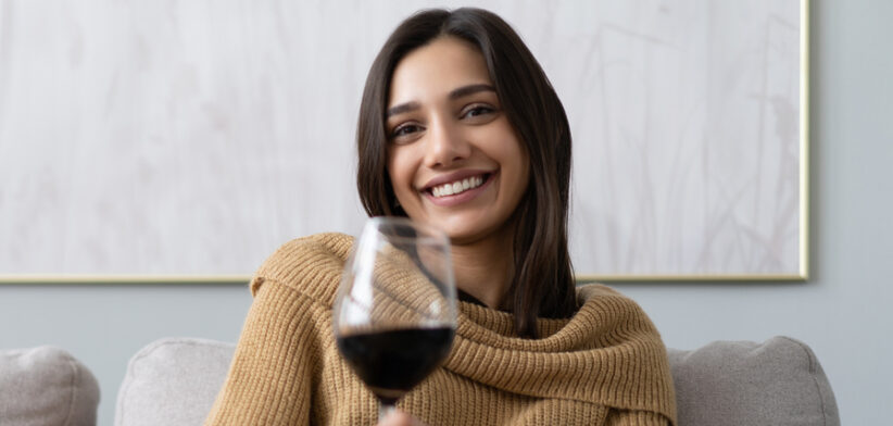 Pretty, attractive, stylish young woman sitting on couch having raised glass with red wine, examine, taste beverage